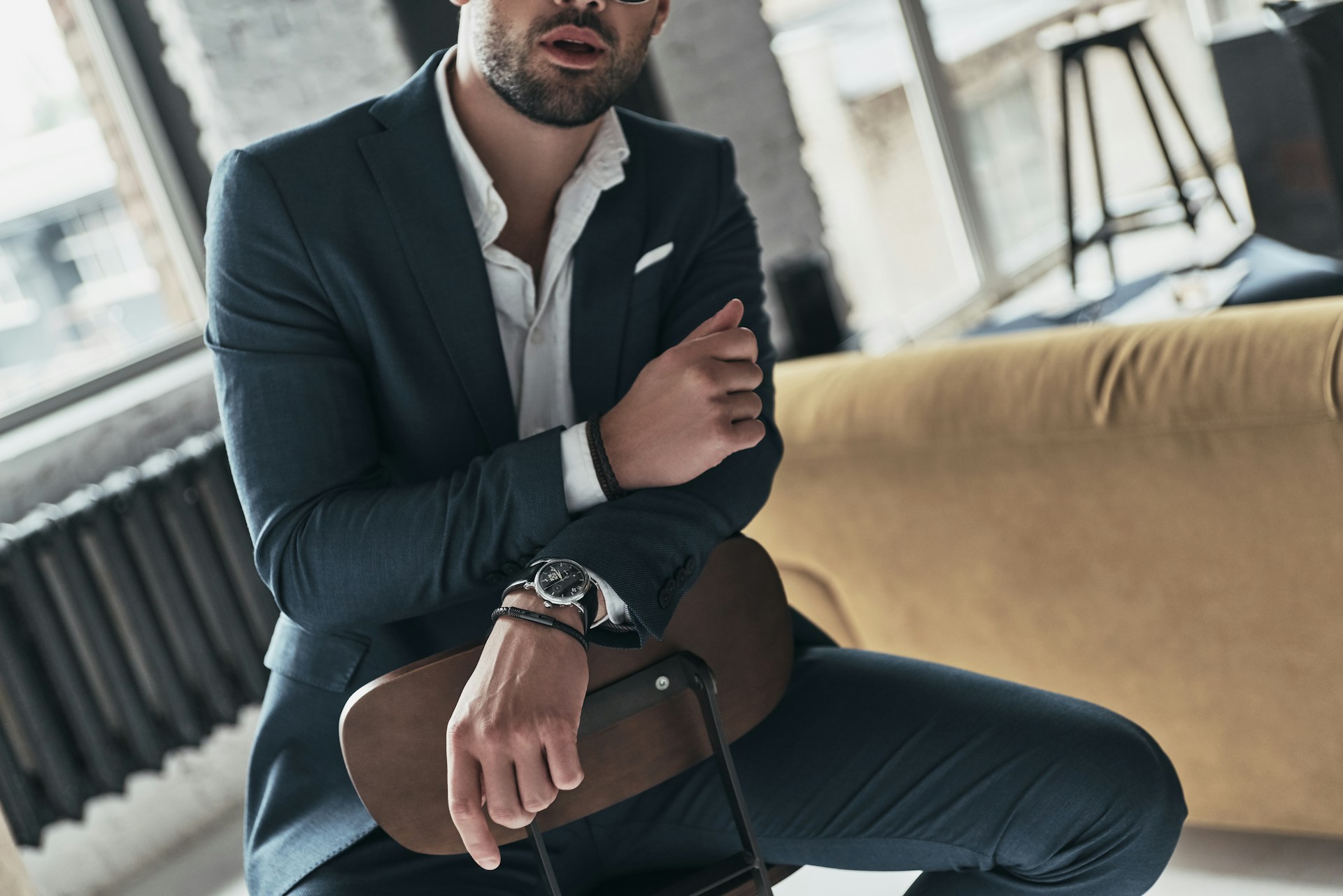 a man in a suit sitting backwards in a chair and wearing a TUDOR watch