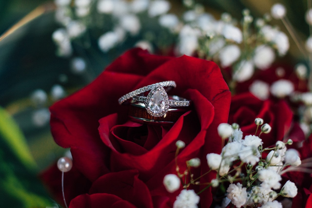 a pear shape halo engagement ring, eternity diamond wedding band, and a man’s white gold and wood grain wedding band poised in the center of a red rose in a bouquet of the flowers.